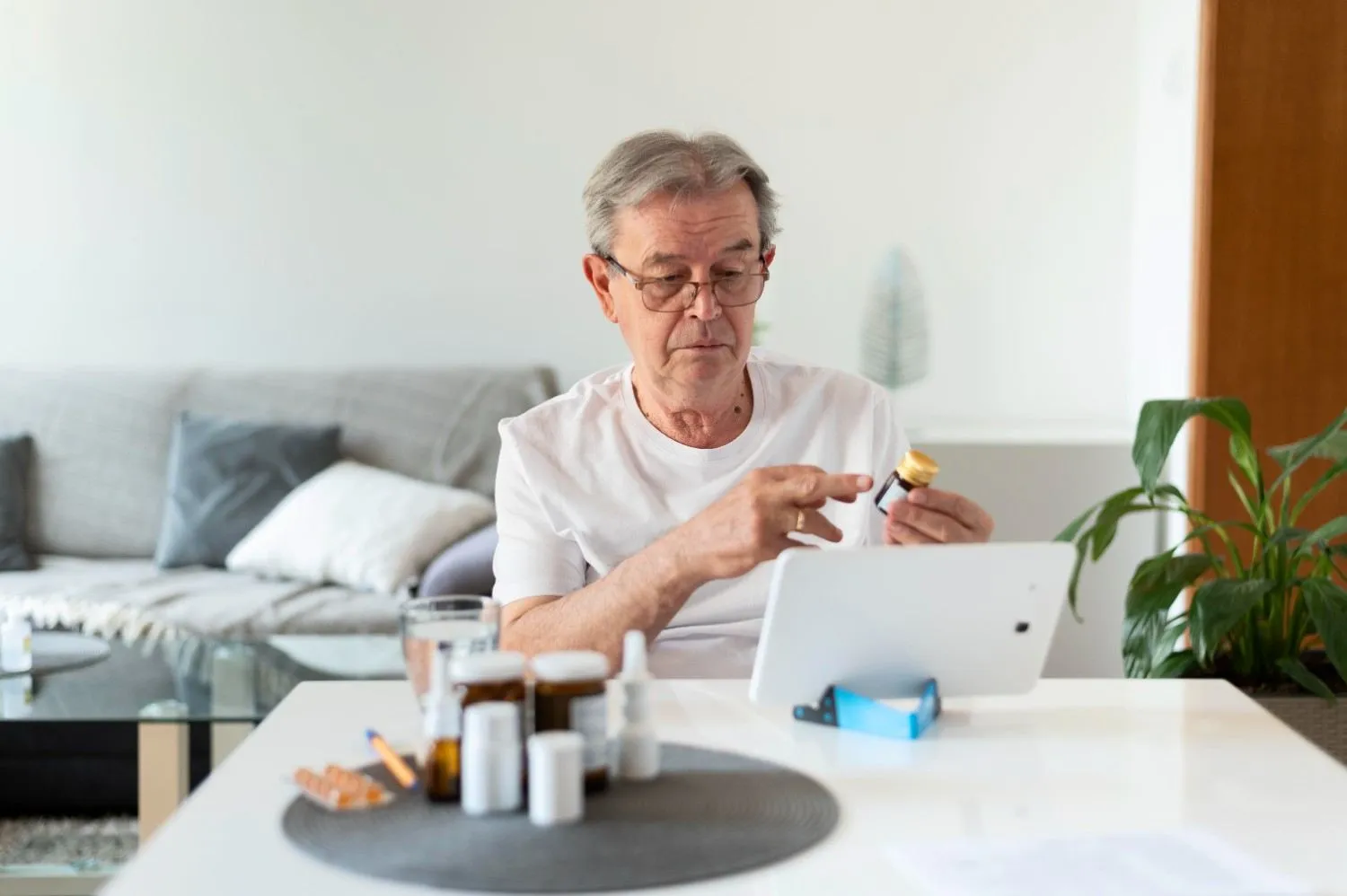 An older man checking a pill bottle against a tablet screen at home